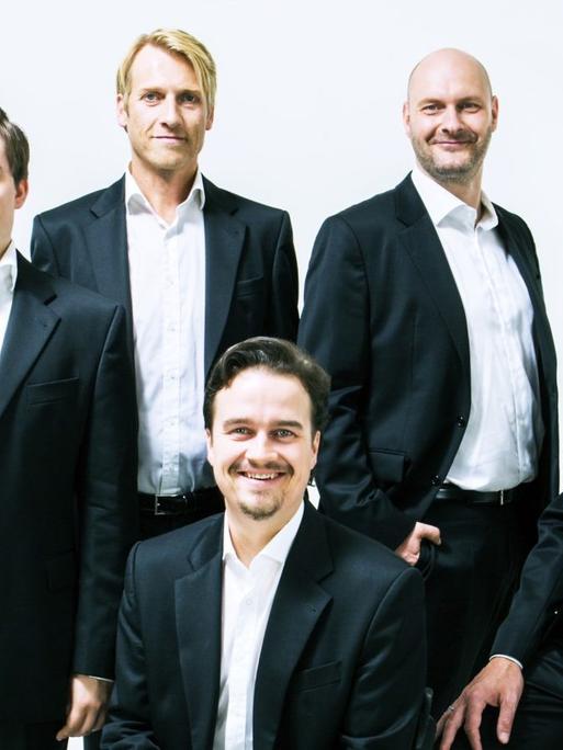Porträt von fünf Männern in schwarzen Anzügen und weißen Hemden: einer sitzt, die anderen stehen hinter ihm.