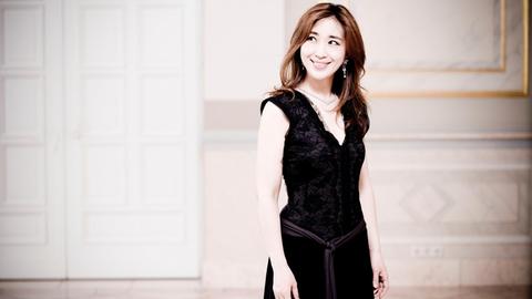 Eine Frau im schwarzen Kleid steht in einem Festsaal und schaut lächelnd zur Seite.