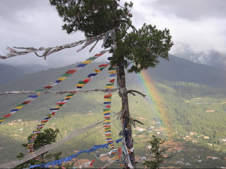 Gebetsfahnen flattern an einem Baum vor einem Regenbogen oberhalb von Thimphu in Bhutan, aufgenommen am 14.07.2013. In dem winzigen Himalaya-Königreich leben viele Menschen noch ein spirituelles Leben. Doch das Land öffnet sich langsam der Außenwelt.
