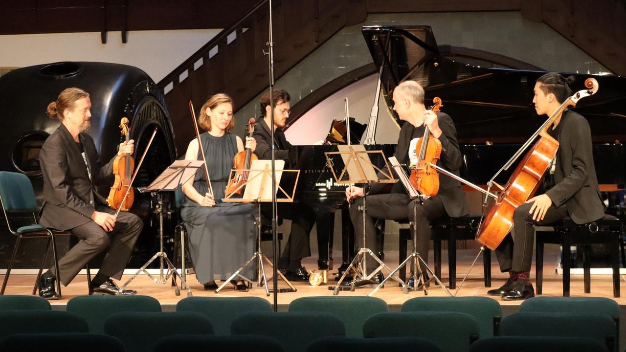 Ein Geiger, eine Geigerin, ein Pianist, ein Bratschist und ein Cellist sitzen in Konzertkleidung mit ihren Instrumenten im Halbkreis auf der Bühne.