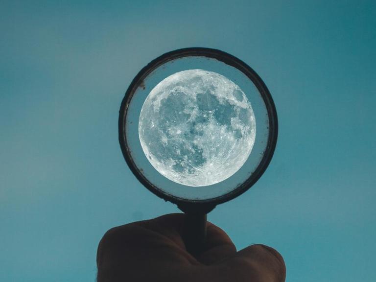 Eine Hand hält einen Kreis gen Abendhimmel, in deren Mitte der Mond leuchtet.