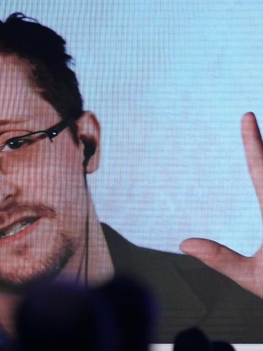 Über verschatteten Sitzreihen scheint ein auf die Bühne projizierter Stream auf, über den Edward Snowden an einer Konferenz teilnimmt.