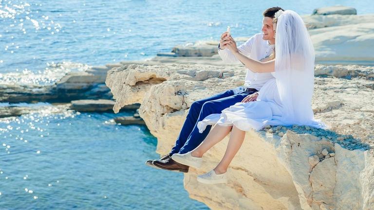 Ein Hochzeitspaar sitzt auf einem Felsen am Strand und macht ein Selfie