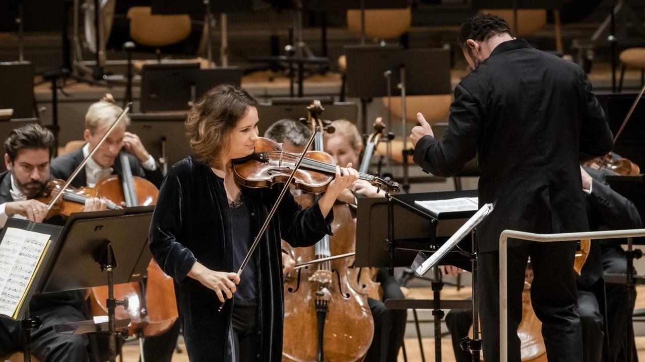 Die Gegerin steht in dunklem Anzug Geige spielend vor den Berliner Philharmonikern.