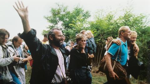 Das Foto zeigt jubelnde DDR-Flüchtlinge, die am 19.8.1989 die Grenze zu Österreich erreichen und eine symbolische Grenzöffnung für die Flucht in den Westen nutzen.