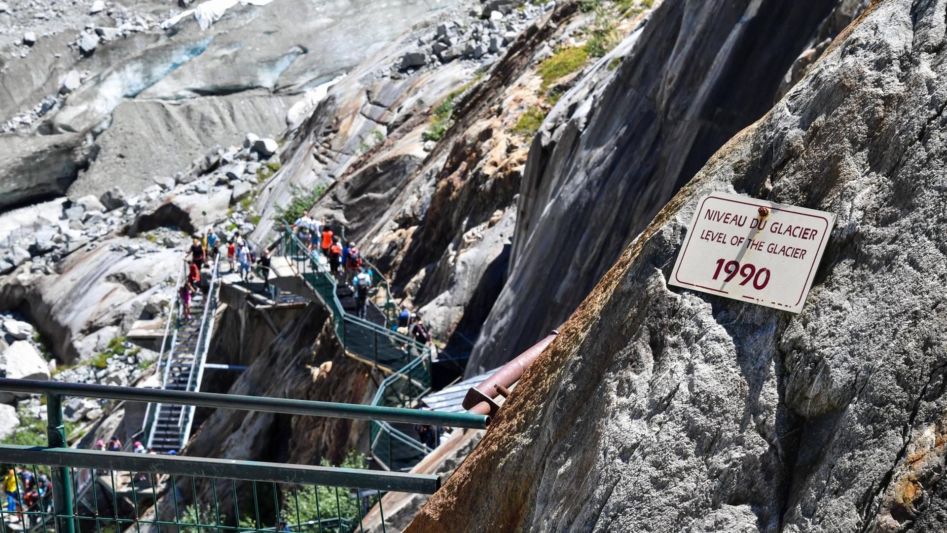 Eine Eisentreppe führt an kahlen, gerölligen Felswänden hinunter. An einem Felsen ist ein Schild angebracht, das darauf hinweist, dass 1990 hier die Decke des Gletschers war.