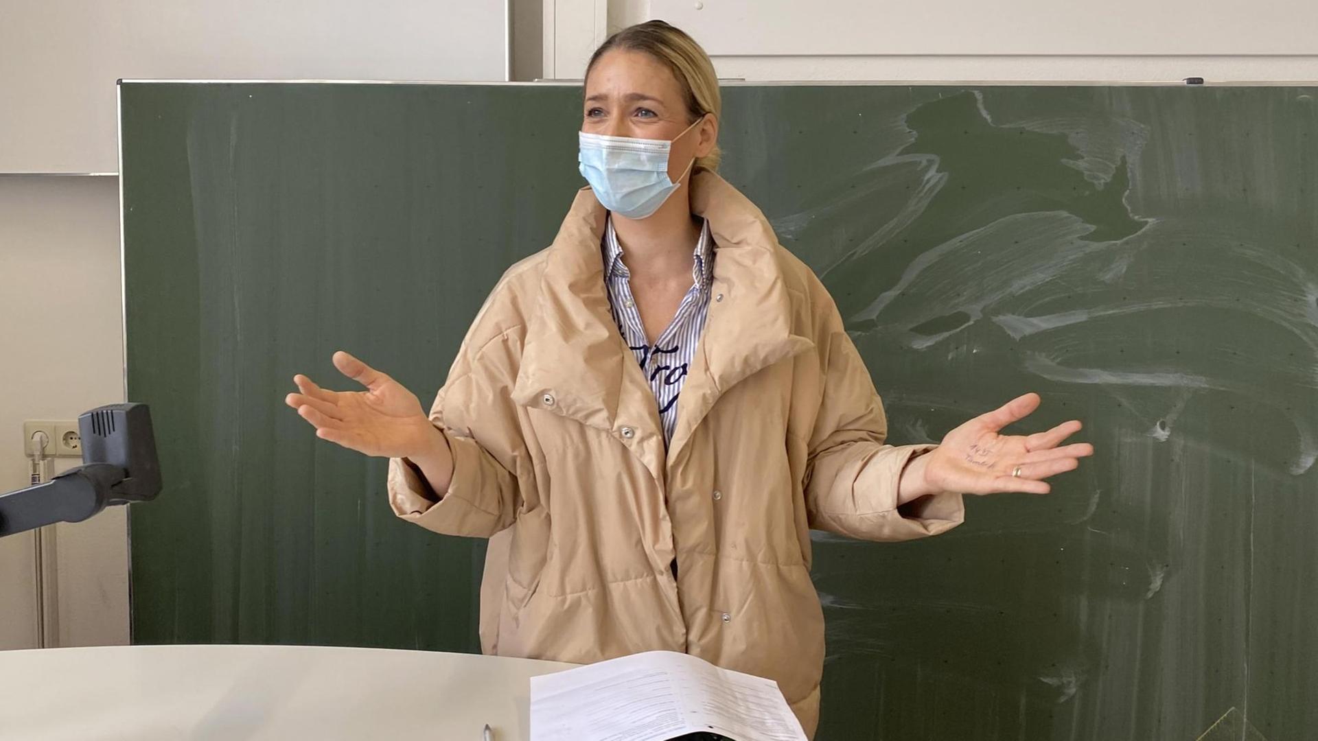 Eine Lehrerin mit Winterjacke und Mundschutz steht vor der Tafel und gestikuliert