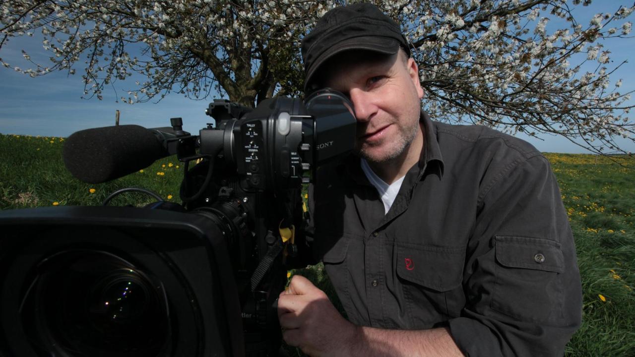 Der Tierfilmer Christoph Hauschild hinter seiner Kamera auf einer grünen Wiese mit einem Baum im Hintergrund