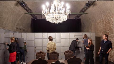 Die Aufnahme zeigt Theatergäste bei der Performance von Tania El Khoury. Sie schließen Türen in einer raumgroßen Tresorwand auf, zwei Theatergäste stecken die Köpfe in die Schließfächer. Die Szenerie wird von einem prächtigen Kronleuchter in der Mitte des Raumes beleuchtet.