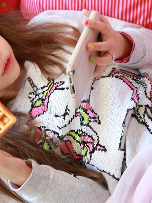 Ein Mädchen liegt auf seinem Bett im Kinderzimmer und schaut auf ein Smartphone (gestellte Szene).