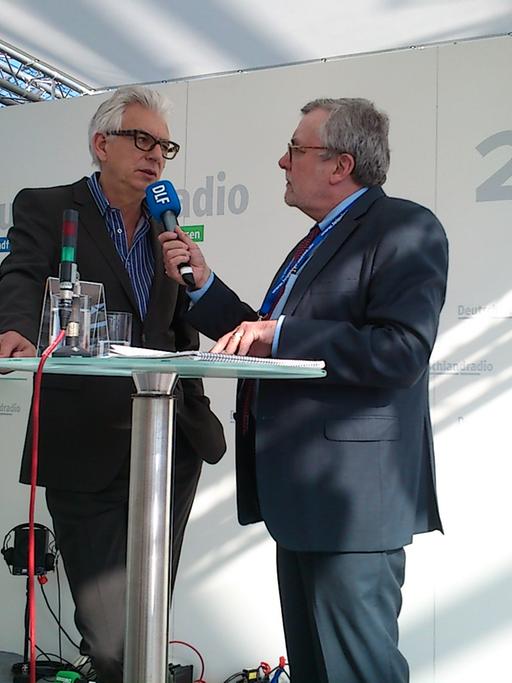 Der "Andruck"-Redakteur Thilo Kößler spricht am Messestand von Deutschlandradio mit dem Buchautor Stephan Wackwitz, beide sind im Halbprofil zu sehen.