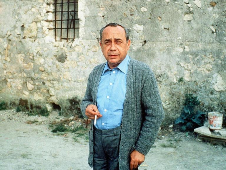 Ein Farbfoto zeigt einen Mann reiferen Alters in grauer Strickjacke mit Stock und Zigarette jeweils in in einer Hand in einem dörflich anmutenden Kontext vor einer Steinmauer