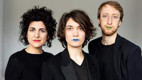 Drei schwarz gekleidete Personen (zwei Frauen und ein Mann) stehen mit farbig geschminkten Lippen nebeneinander.