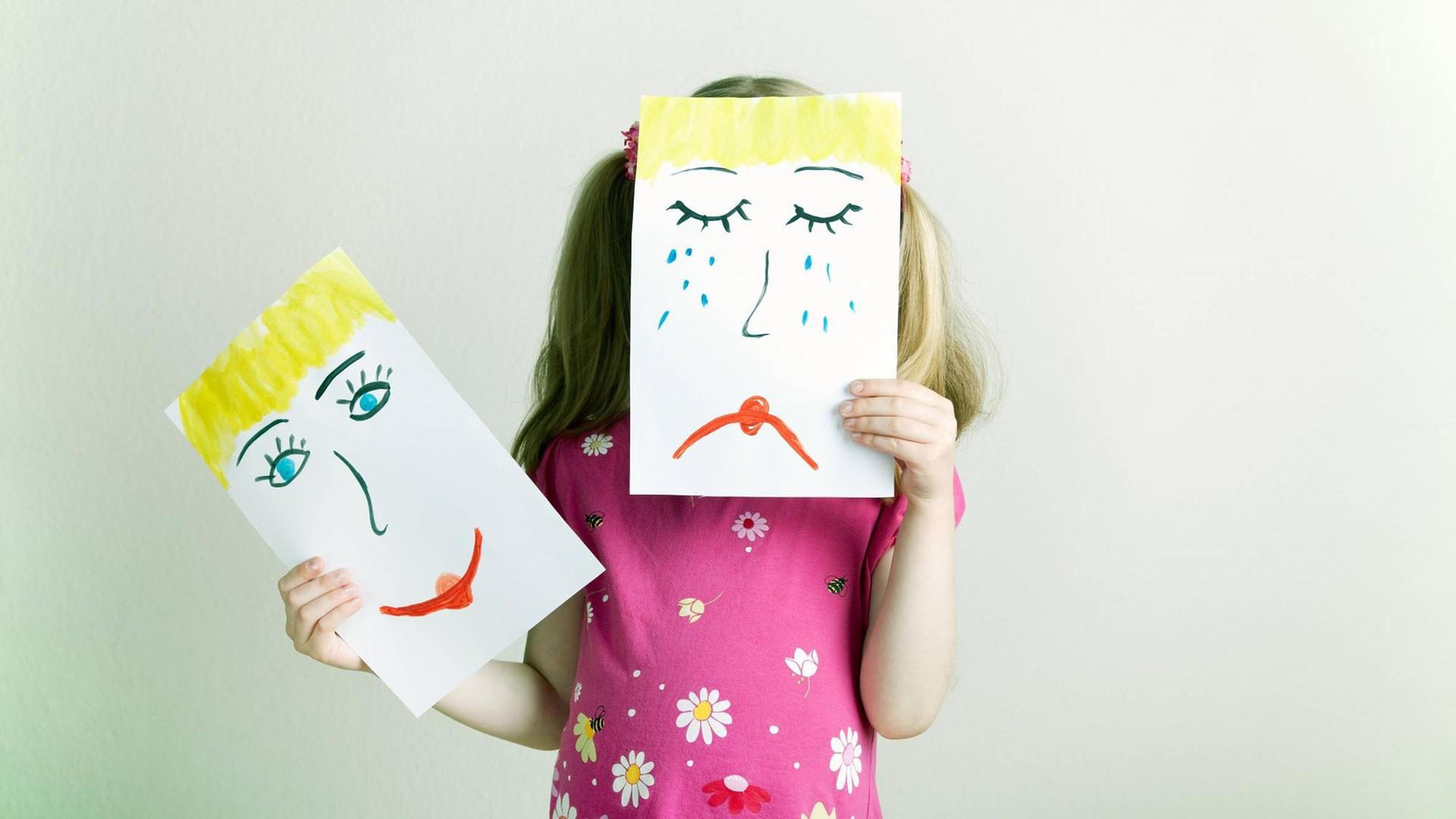 Ein Mädchen hält Zeichnungen mit einem lachendem und einem weinenden Gesicht in die Höhe.