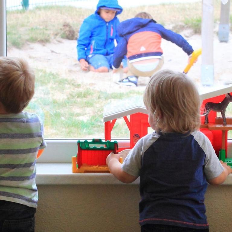 Zwei Kinder stehen am Fenster einer Kita und gucken anderen Kindern draußen beim Spielen zun