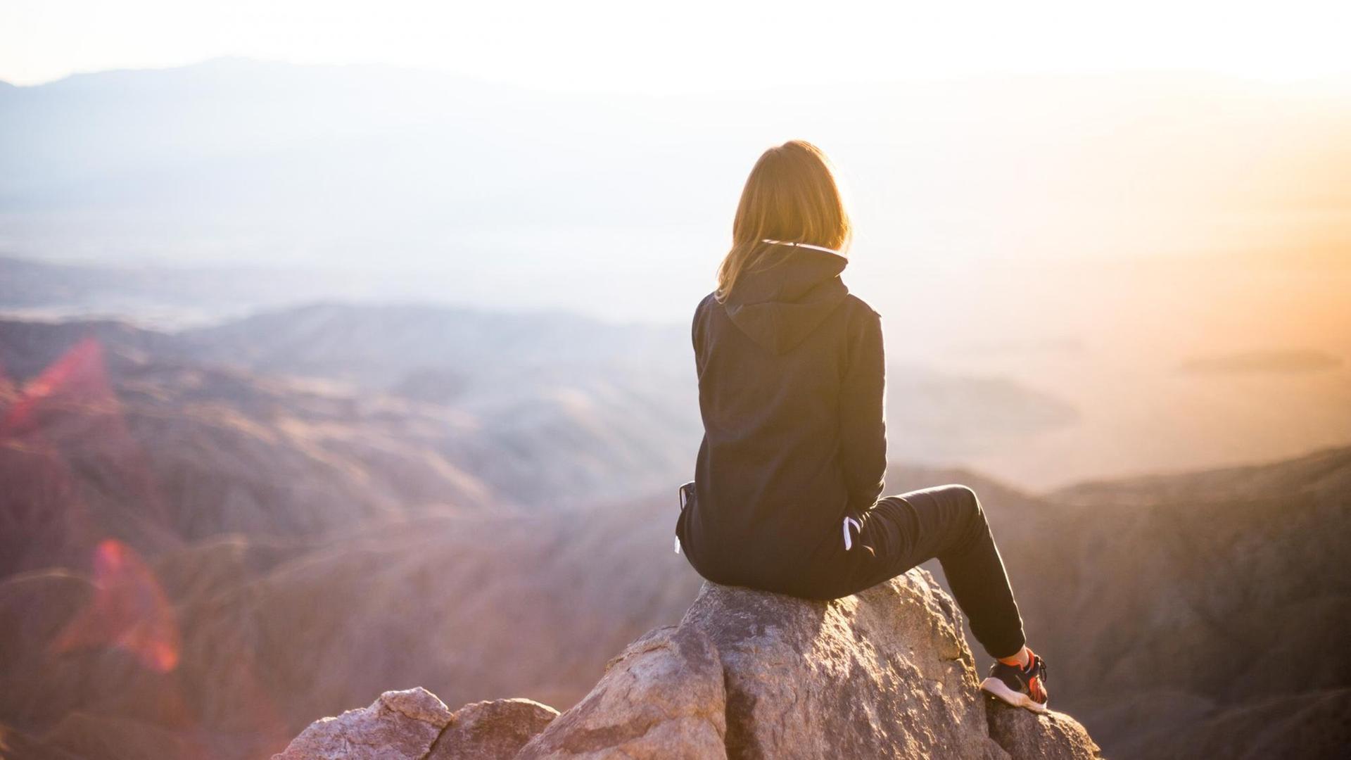 Eine Frau sitzt auf dem Gipfel eines Berges, den Rücken zur Kamera.