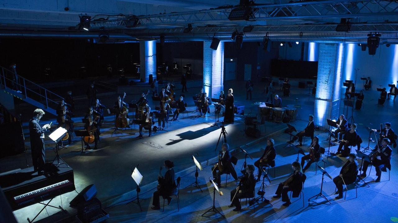 In einem blau ausgeleuchteten Industrieraum sitzt verstreut ein Orchester, das eine Sängerin weiträumig umringt samt Dirigenten, der auf einem erhöhten Pult steht.