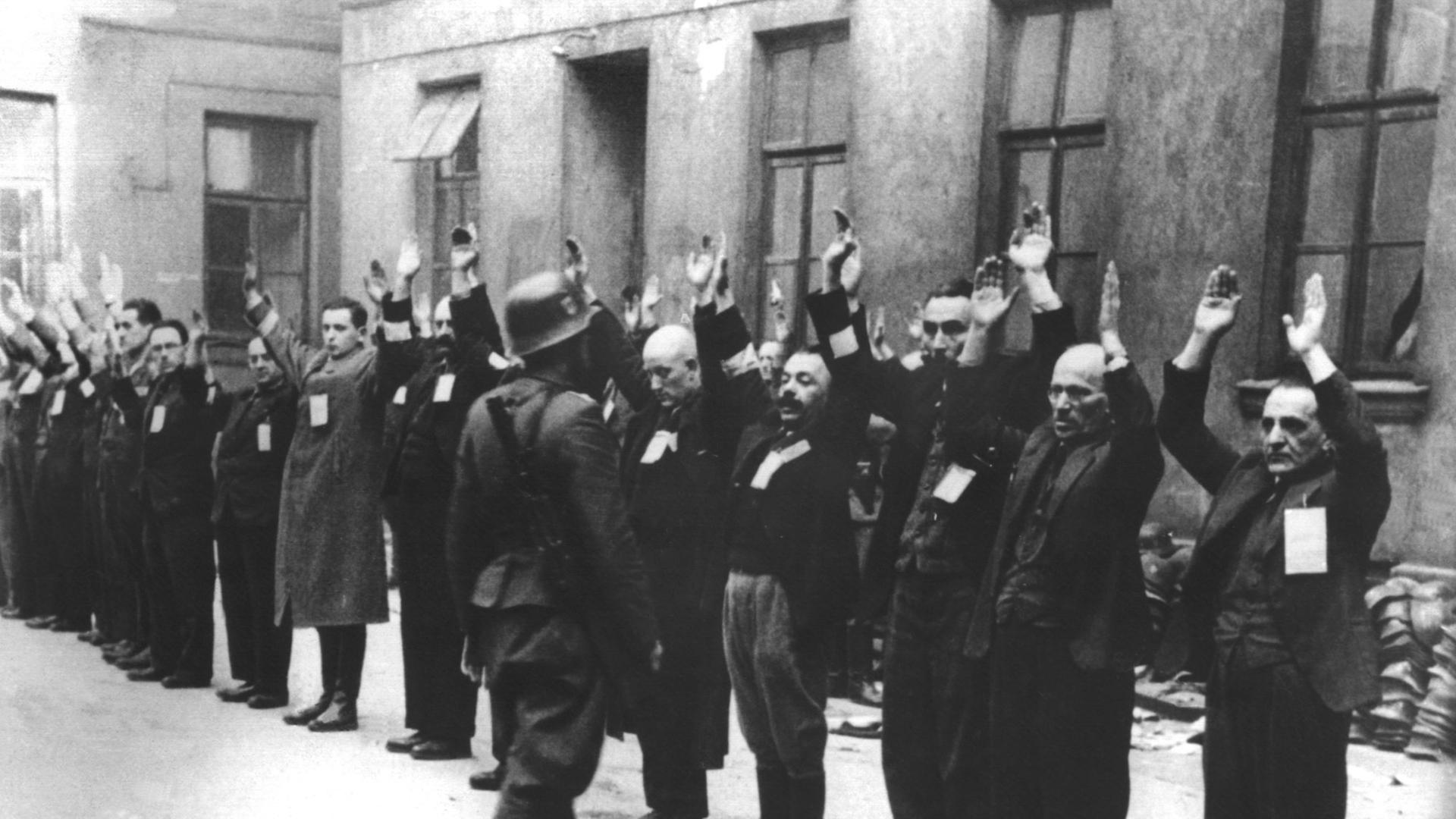 Bewacht von einem deutschen Soldaten stehen Bewohner des Warschauer Ghettos mit erhobenen Armen in einem Innenhof.