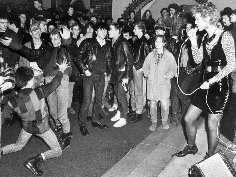 Auf der schwarzweiss Aufnahme ist das Benefizkonzert in der Berliner Erlöserkirche 1988 zu sehen. Eine Sängerin steht vor einer tanzenden Masse aus jungen Menschen.