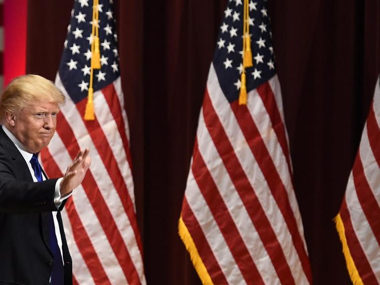 Man sieht Donald Trump, der winkt, im Hintergrund drei große US-Fahnen.