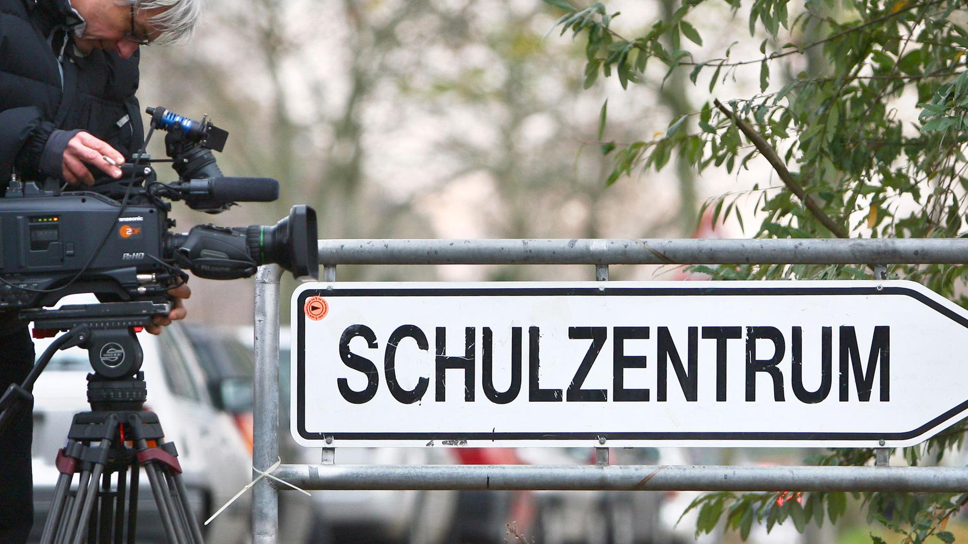 Ein Kameramann richtet seine Kamera auf ein Schild, das den Weg Richtung "Schulzentrum" weist.