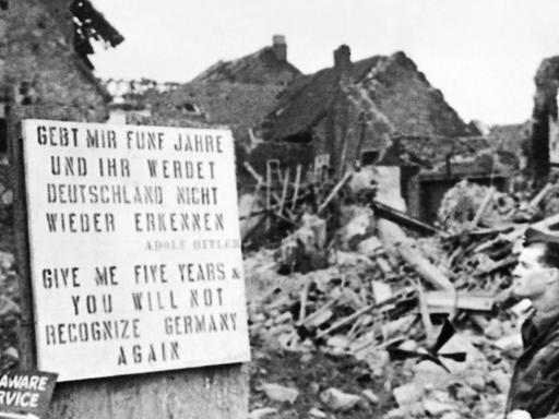 "Gebt mir fünf Jahre und ihr werdet Deutschland nicht wiedererkennen" steht in Deutsch und Englisch auf einem Schild vor einem Trümmerberg, ein Mann liest das Schild, schwarz-weiß-Aufnahme.