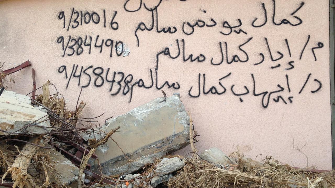 Arabische Schriftzeichen und Zahlen stehen auf auf einer Hauswand, davor liegen Steine und Trümmer