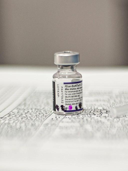 Ein Fläschchen mit Impfstoff und eine leere Spritze liegen auf einem bedruckten Blatt Papier.