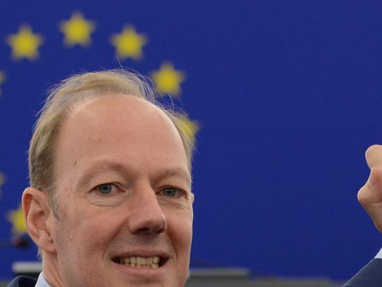 Journalist, Satiriker und Mitglied des Europäischen Parlaments, Martin Sonneborn, macht eine Siegerpose mit den Händen, dabei leuchten die Sterne der EU-Flagge über seinem Kopf