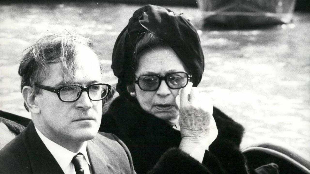 Ein Mann und eine Frau sitzen in einer Gondel. Sie trägt einen schwarzen Hut und hält ein Taschentuch gegen ihre Wange.