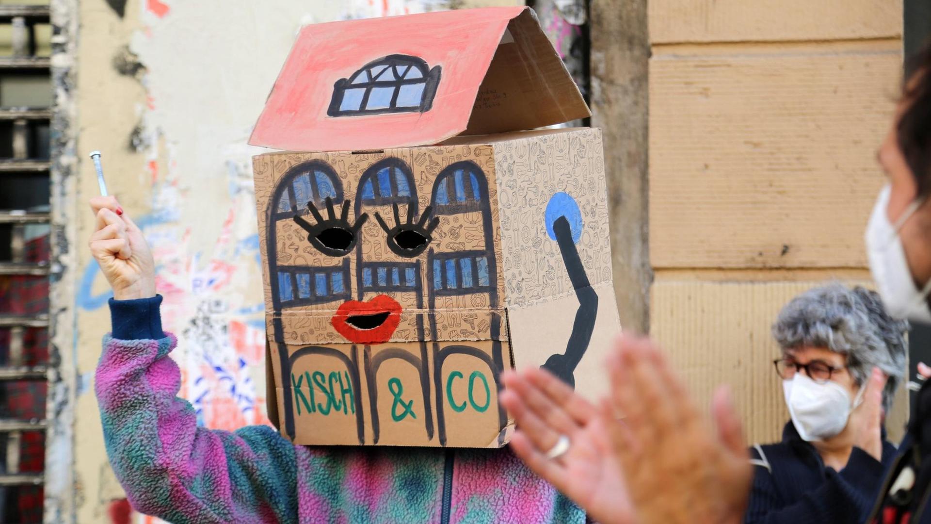 Ein Demonstrant hat sich einen zum Haus umgestalteten Pappkarton auf die Schultern und über seinen Kopf gesetzt. Der Pappkarton stellt das Haus dar, in dem sich die Räume von Kisch & Co. bislang befanden