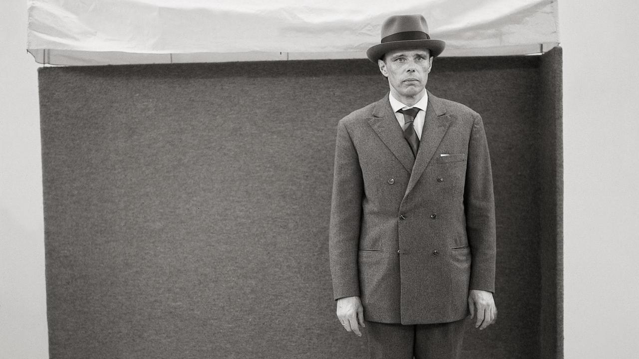 Joseph Beuys steht mit Hut und Anzug vor einer Skulptur.