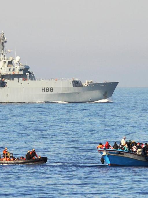 Flüchtlinge werden im Mittelmeer von Bundeswehrsoldaten auf zwei Speedbooten von ihrem Holzboot abgeholt und anschließend zur Fregatte Schleswig-Holstein gebracht. Im Hintergrund ist die britische HMS Enterprise zu sehen.