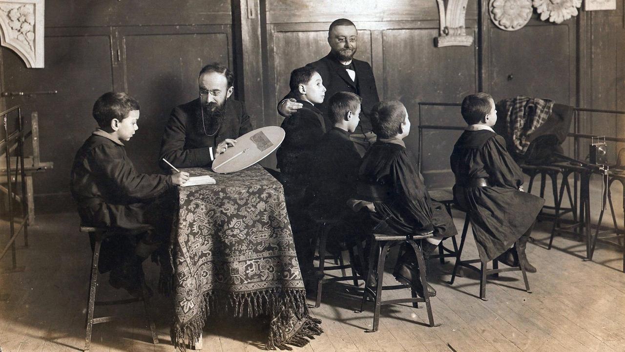 Schwarzweißaufnahme von 1907: In einem dunkelgetäfelten Schulzimmer sitzen mehrere Jungen auf Holzstühlen. Der Psychologe Alfred Binet mit schwarzem Frack, Brille und Bart zeigt ihnen Aufgaben aus einem psychologischen Test.