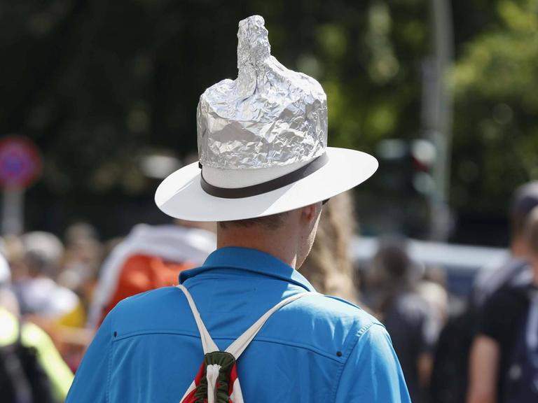 Ein Mann mit einem Aluhut steht mit dem Rücken zum Betrachter am Rand einer Demonstration