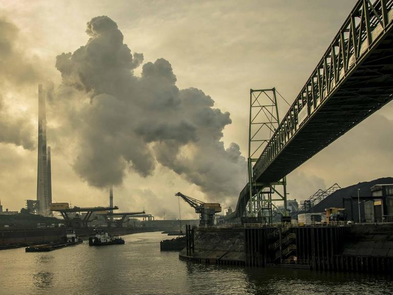 Eine graue Industrielandschaft am Rhein. An den Ufern des Flusses stehen Stahlwerke der Firma Thyssenkrupp. Aus Fabrikschloten steigt Rauch in den Himmel.