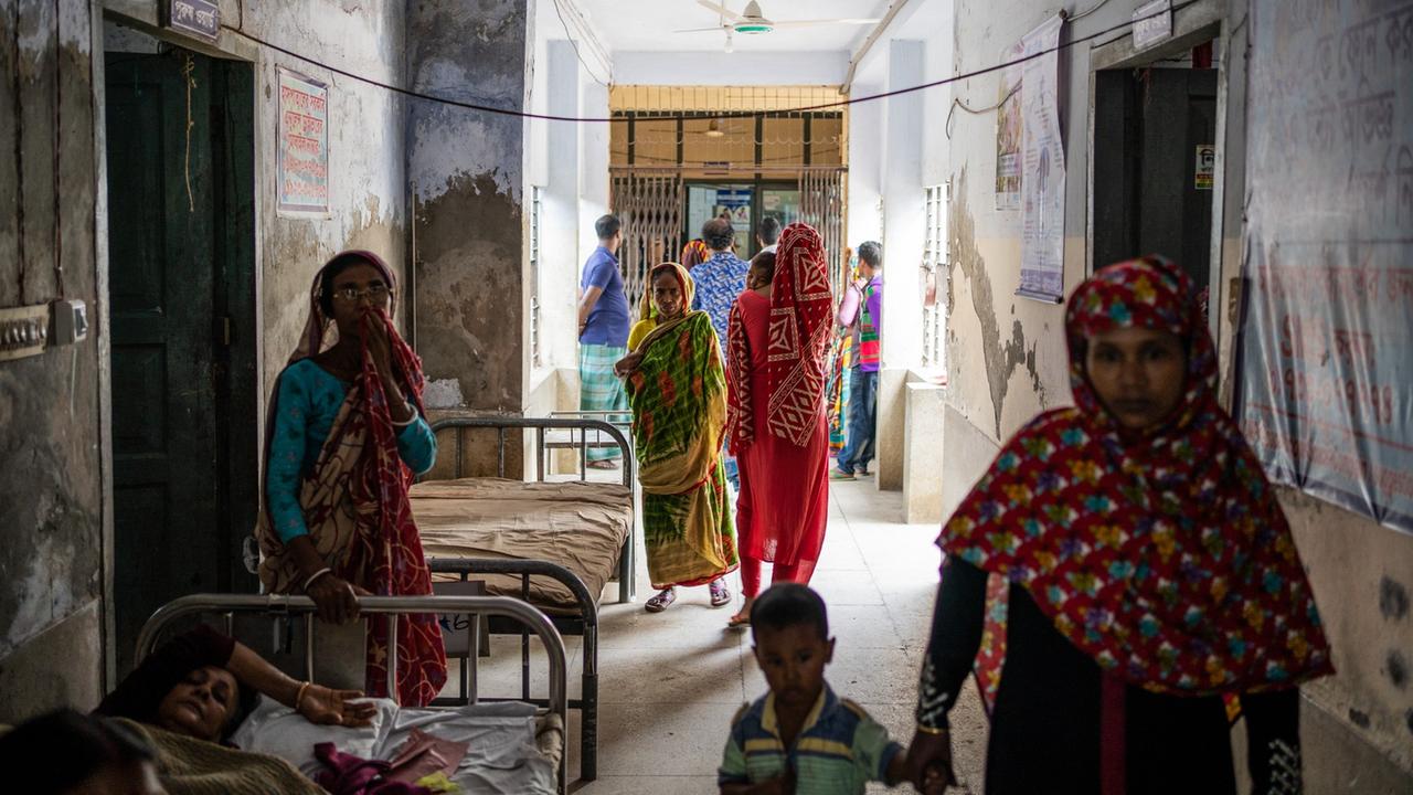 Ein Gang im Krankenhaus in Khulna, links liegt eine Patientin im Bett, rechts geht eine Mutter mit Kind an der Hand. Im Hintergrund wartende Angehörige