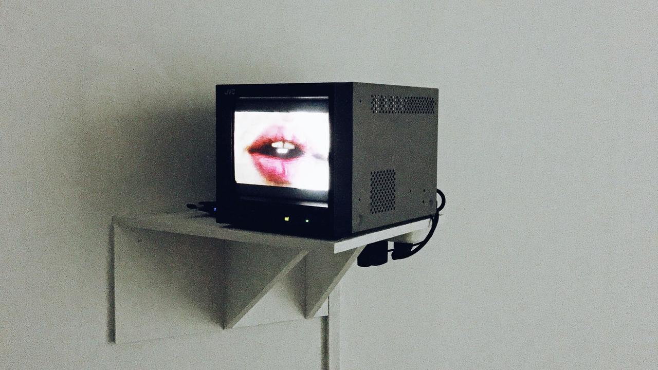 Auf einem Monitor vor einer grauen Wand erscheint ein roter Kussmund.