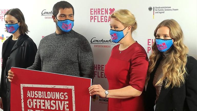 Vier Personen stehen mit Mundschutz vor einer Plakatwand. Zwei Menschen halten ein Schild, auf dem steht "Ausbildungsoffensive Pflege".