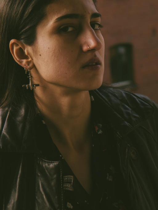 Leyla Yenirce mit Nasenring und Flugzeug-Ohrring trägt Lederjacke und schaut in die Kamera. Im Hintergrund Backsteinarchitecktur.