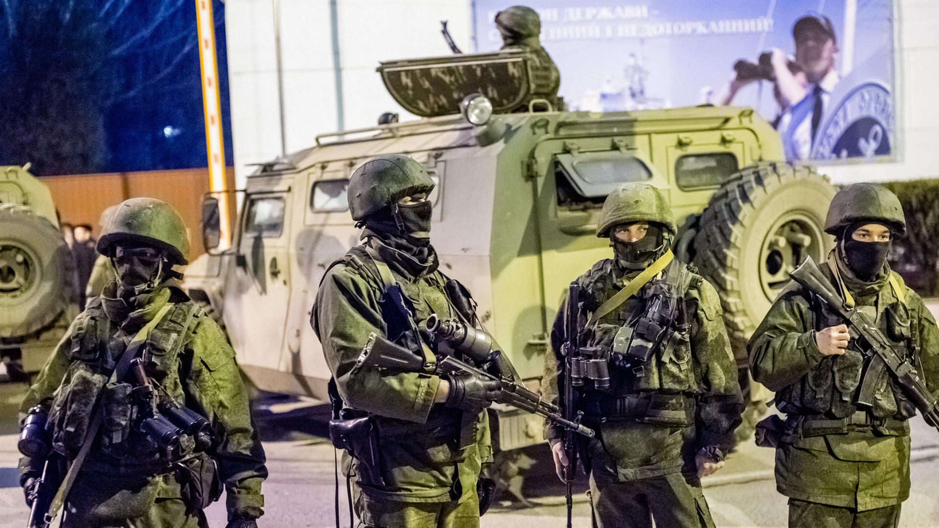 Mehrere vermummte Soldaten in grünen Uniformen mit Helmen und Maschinengewehren stehen vor einem Panzer