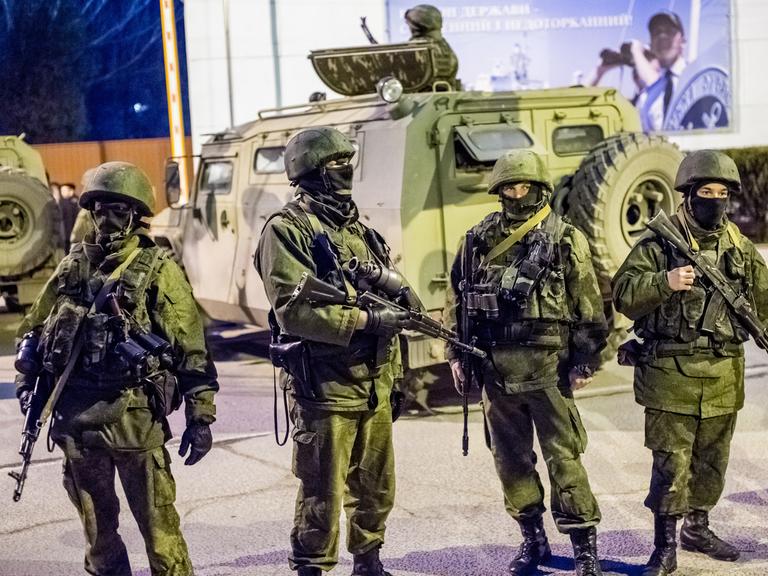 Mehrere vermummte Soldaten in grünen Uniformen mit Helmen und Maschinengewehren stehen vor einem Panzer