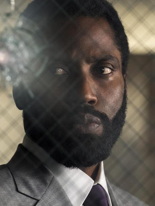 Der Hauptdarsteller John David Washington im neuen Christopher Nolan Film "Tenet". Er steht hinter einer von Kugeln getroffenen Glasscheibe.