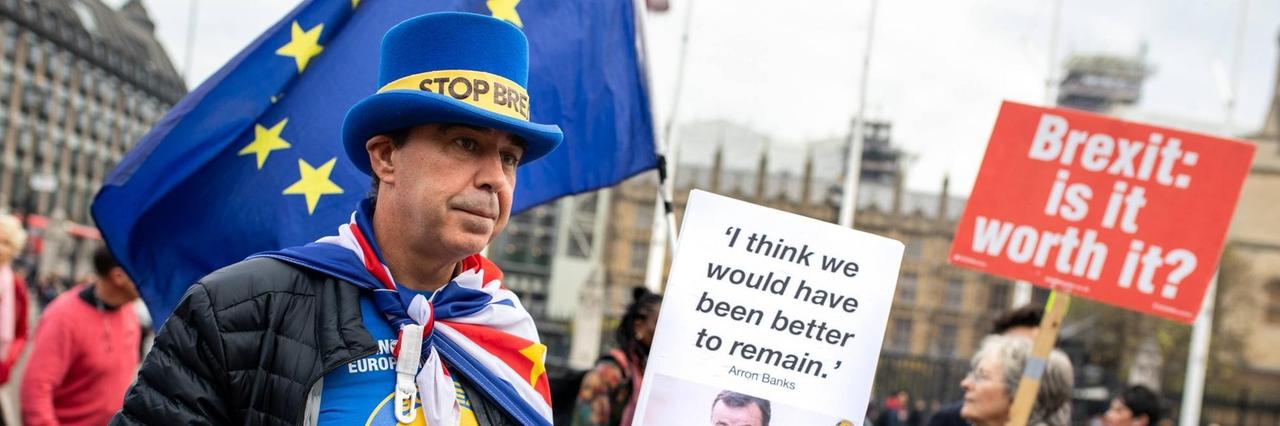 
Ein Mann demonstriert mit anderen Brexit-Gegnern auf dem Londoner Parliament Square.  Er trägt einen blauen Zylinder mit der Aufschrift: "Stop Brexit".  Außerdem hält er ein Plakat mit einem Zitat von Arron Banks hoch: "I think we would have been better to remain." Im Hintergrund ist eine Frau zu sehen, die ein Plakat mit der Aufschrift hochhält: "Brexit: is it worth it?"
