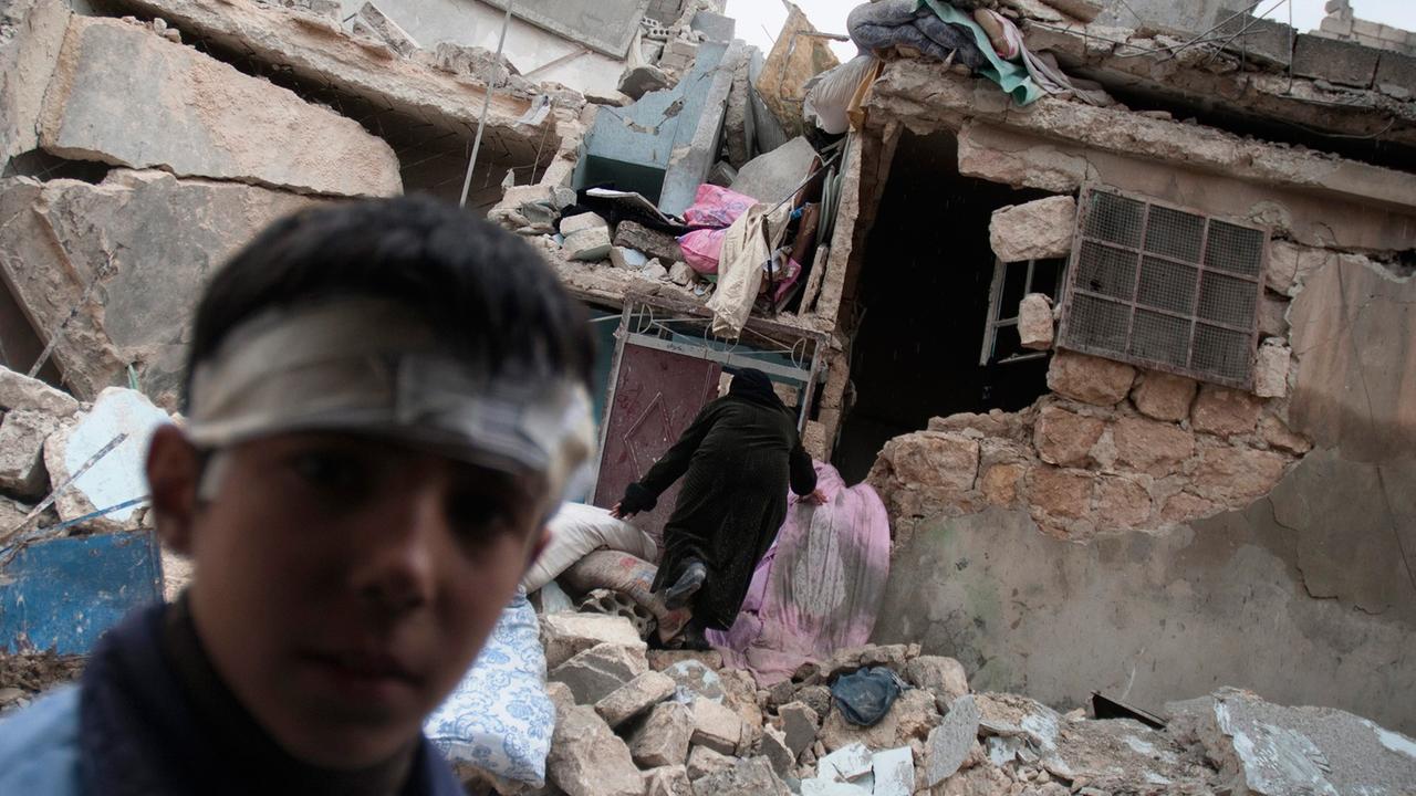 Im Hintergrund sind Trümmer eines zerstörten Hauses zu sehen, durch die ein Mann mit dem Rücken zur Kamera klettert. Im Vordergrund schaut ein etwa zehnjähriger Junge mit einem Kopfverband Richtung Kamera.