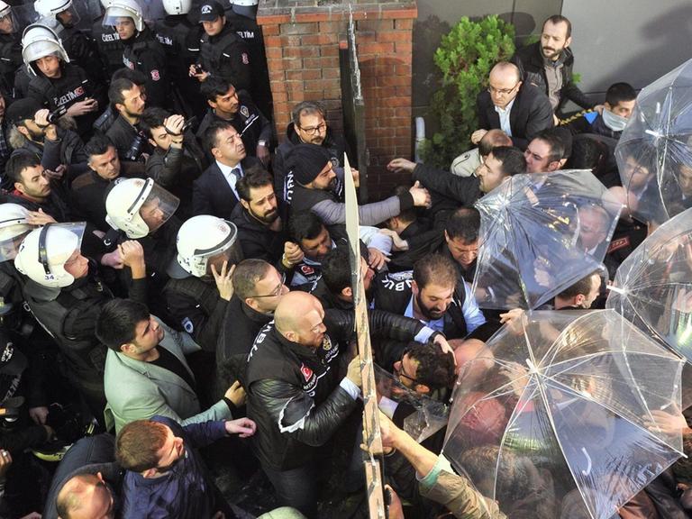 Man sieht von oben, wie die Polizei und ihre Gegner rechts bzw. links eines Tores stehen. Einige der Gegner haben Schirme aufgespannt, die sie in Richtung der Polizisten halten.