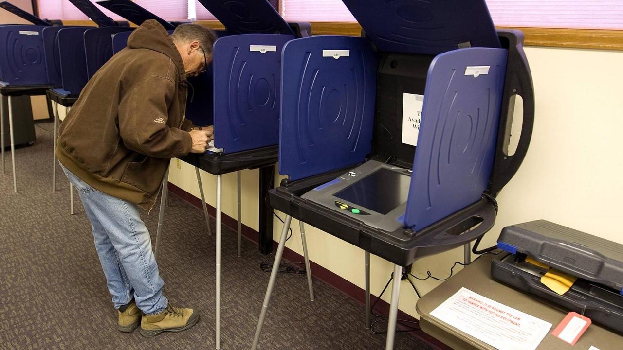 Mehrere Wahlcomputer mit Sichtschutz an den Seiten und oben stehen nebeneinander in einem Wahllokal an der Wand. Ein Mann beugt sich über einen der Computer.