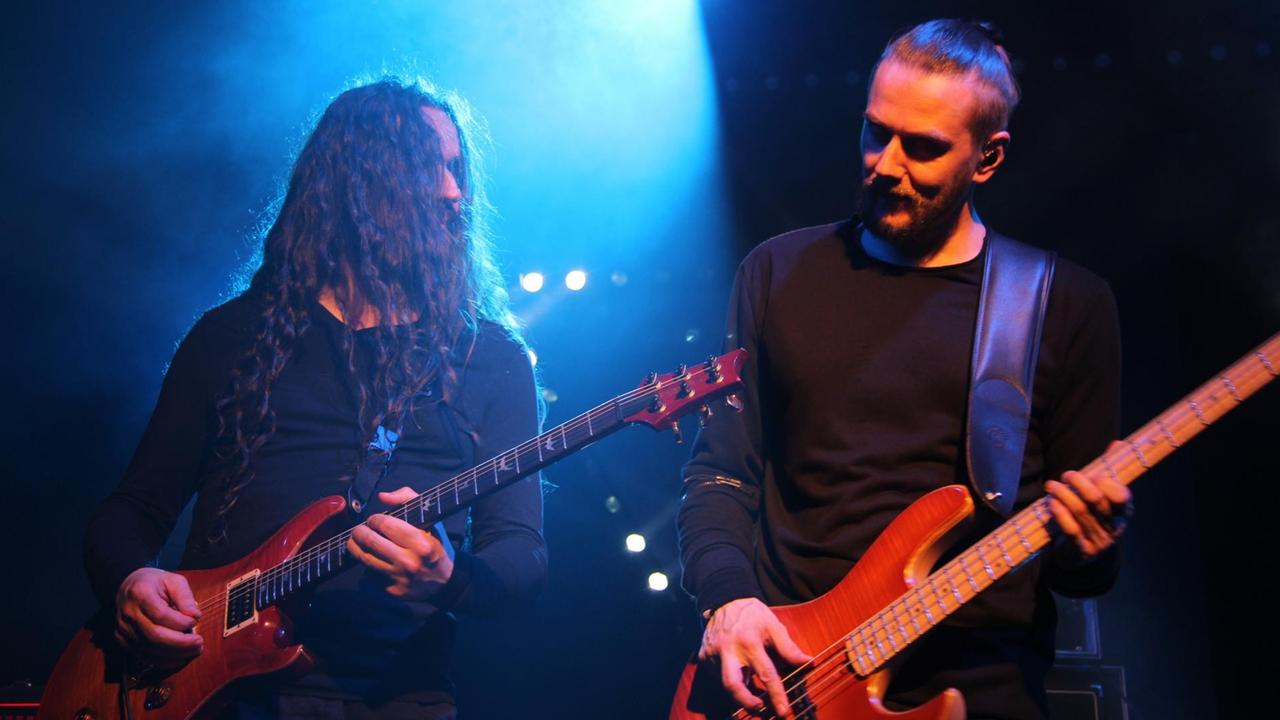 Zwei Männer stehen auf der Bühne, einer am Bass, einer mit Gitarre. Die Bühne ist in blaues Licht getaucht.