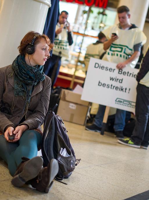 Eine junge Frau mit Kopfhörern wartet am 15.10.2014 im Hauptbahnhof Hannover (Niedersachsen) sitzend auf einen Zug, während im Hintergrund Lokführer stehen.