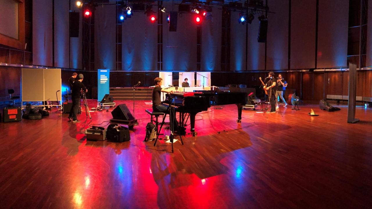 Fünf Musiker stehen in einem grossen Saal und machen Musik. Im Vordergrund sitzt ein Mann am Klavier.